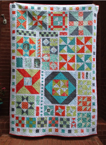 Start here: A colorful patchwork quilt hangs against a brick wall, featuring various geometric patterns in shades of orange, teal, green, white, and gray with swirled stitching throughout.