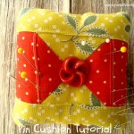A square pin cushion with yellow and red patchwork fabric, inspired by a classic quilt block design, is decorated with a red button in the center. Several pins rest atop it on a wooden surface. Text at the bottom reads "Pin Cushion Tutorial.