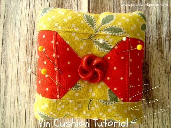 A square pin cushion with yellow and red patchwork fabric, inspired by a classic quilt block design, is decorated with a red button in the center. Several pins rest atop it on a wooden surface. Text at the bottom reads "Pin Cushion Tutorial.