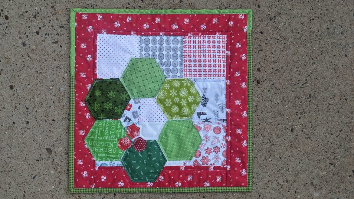 A small Hexagon Quilt with a red border, green striped edge, and a patchwork center. Large hexagons in various green prints overlay squares of white, gray, and red. The quilt is displayed on a concrete surface.