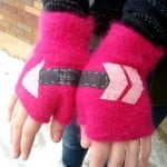 Two hands in bright pink gloves with arrow-patterned fabric patches point left. The person stands outdoors, near snow and a brick wall.