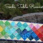 A colorful quilted table runner with a diamond pattern in rainbow colors, inspired by the Sochi Olympics, is displayed on a metal table outdoors with evergreen branches in the background. Text reads "Sochi Table Runner.
