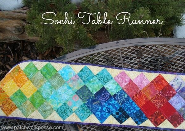 25+ Easy Sewing Projects to Brighten Your Kitchen 15 A colorful quilted table runner with a diamond pattern in rainbow colors, inspired by the Sochi Olympics, is displayed on a metal table outdoors with evergreen branches in the background. Text reads "Sochi Table Runner.