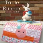 A colorful Easter table runner with a bunny face design drapes over a table. On top sits a ceramic white bunny statue holding a blue egg, in front of a brown and red brick wall. Text reads: "Easter Table Runner.