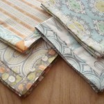Four neatly folded pieces of fabric with various pastel-colored patterns, including stripes, florals, and geometric designs, arranged on a wooden surface—perfect for your next Sewing Tutorial or self binding napkin project.