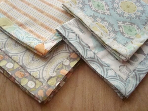 25+ Easy Sewing Projects to Brighten Your Kitchen 7 Four neatly folded pieces of fabric with various pastel-colored patterns, including stripes, florals, and geometric designs, arranged on a wooden surface—perfect for your next Sewing Tutorial or self binding napkin project.