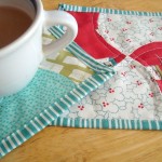 A cup of coffee sits on a wooden table atop vibrant quilted coasters, lovingly crafted from a cherished fabric stash. The coasters boast red, teal, and patterned designs with striped borders, enhancing their patchwork charm.