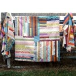 Three colorful patchwork quilts, including a striking Jelly Roll Quilt, hang on a wooden fence outdoors. Their vibrant patterns and stripes pop against the rustic background, creating a charming scene perfect for any simple quilt enthusiast.