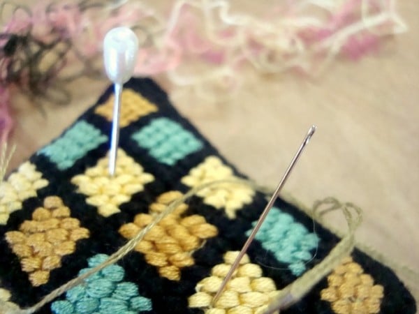 Close-up of a pin cushion with a pearl-headed pin and a needle threaded with green thread, resting on a vintage embroidery pattern of yellow and blue squares. Tangled threads add texture in the background.