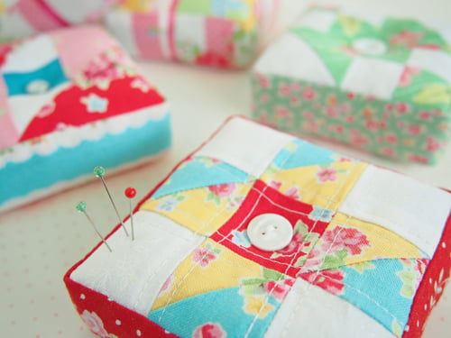Colorful, square pincushions made from patchwork fabric are arranged on a light surface. One pincushion in the foreground, inspired by free pincushion patterns, features three pins and a white button in the center.