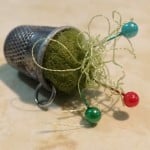 A metal Thimble Pin Cushion filled with green felt and tangled thread, with three straight pins—one blue, two red—stuck in the felt. Perfect for quilting, it rests on a light, textured surface.