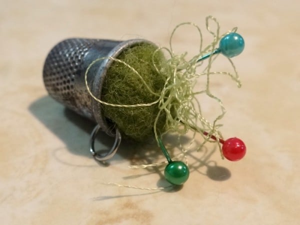 A metal Thimble Pin Cushion filled with green felt and tangled thread, with three straight pins&mdash;one blue, two red&mdash;stuck in the felt. Perfect for quilting, it rests on a light, textured surface.