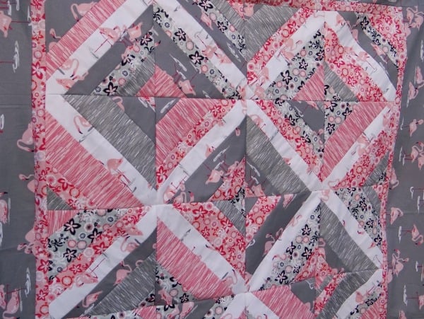 25 Must Sew Scrappy Strip Quilt Patterns 7 A quilt with a geometric pattern in pink, white, black, and gray fabrics, featuring string pieced sections and prints like stripes, florals, and flamingos arranged in diagonal and triangular shapes.