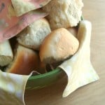 A green bowl lined with a patterned Bread Basket Cover contains several pieces of golden brown dinner rolls, with soft, fluffy textures visible. The bowl is placed on a light wooden surface.
