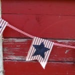 A string of triangular fabric flags crafted from a vibrant fabric stash hangs against a weathered red wooden wall. Each flag features a blue star on red and white stripes, connected by a red and white striped string.