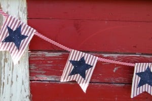 A string of triangular fabric flags crafted from a vibrant fabric stash hangs against a weathered red wooden wall. Each flag features a blue star on red and white stripes, connected by a red and white striped string.