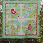 A colorful quilt with four large blocks featuring early bird appliqué in red, patchwork patterns in green, blue, and yellow, and a floral border, hanging outdoors among green bushes.