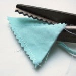 A pair of pinking shears cutting light blue fabric for fabric bunting, creating a zigzag edge. The fabric is partially cut, with visible stitches and loose threads along the edge—a faster method to make fabric bunting projects.