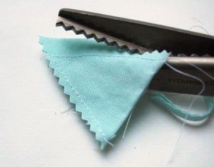 A pair of pinking shears cutting light blue fabric for fabric bunting, creating a zigzag edge. The fabric is partially cut, with visible stitches and loose threads along the edge&mdash;a faster method to make fabric bunting projects.