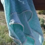 A light blue double gauze baby blanket with white trim and fluffy pom poms draped over a wooden railing outdoors, with shrubs and a clear sky in the background.