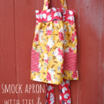 A colorful floral apron smock with red and yellow patterns, featuring two striped pockets and ties, hangs on a red wooden wall. Text reads: "Smock apron with ties & pockets.