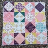 A colorful patchwork Layer Cake Quilt featuring square and diamond patterns in pink, yellow, purple, green, and white prints is laid out on a gravel surface.