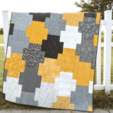 A patchwork quilt with interlocking rectangles in yellow, white, black, and various shades of gray illustrates lap quilt patterns using fat quarters, draped over a white fence outdoors with grass and trees in the background.