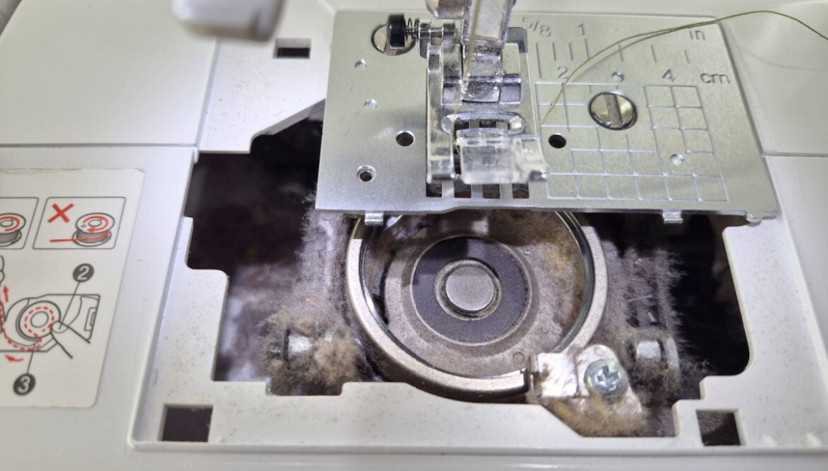 Sewing Machine Maintenance Tracker – Free Printable 1 Close-up of a sewing machine bobbin area filled with dust and lint, with the metal bobbin case exposed and sewing instructions visible on the left—highlighting just how useful a machine cleaning tracker can be for maintenance.