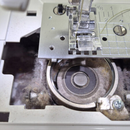 Close-up of a sewing machine bobbin area filled with dust and lint, with the metal bobbin case exposed and sewing instructions visible on the left—highlighting just how useful a machine cleaning tracker can be for maintenance.