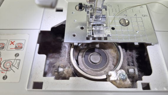Close-up of a sewing machine bobbin area filled with dust and lint, with the metal bobbin case exposed and sewing instructions visible on the left—highlighting just how useful a machine cleaning tracker can be for maintenance.