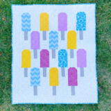 A colorful quilt featuring yellow, purple, and blue popsicles with patterned designs on a white background, laid out on green grass—perfect inspiration for easy baby quilts.