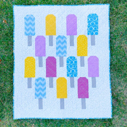 A colorful quilt featuring yellow, purple, and blue popsicles with patterned designs on a white background, laid out on green grass&mdash;perfect inspiration for easy baby quilts.