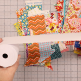 A person holds a roll of white paper tape, unspooling a strip above sewing scraps on a gridded cutting mat. A green ruler is partially visible in the corner, suggesting creative sewing techniques.