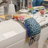 A colorful quilted pincushion brimming with pins is attached to a white sewing machine in a cluttered sewing workspace.