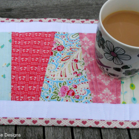 A patchwork quilted placemat, enhanced with vibrant mug rug patterns, features colorful fabric strips in pink, red, and blue. A floral-designed mug filled with tea or coffee rests on it. The placemat's edges charm with a heart pattern, showcasing the art of quilting.