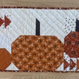 A patchwork quilted table runner showcases three stylized pumpkins in rich shades of orange and brown, framed by decorative geometric patterns and triangles. The backdrop is a crisp white with an elegant brown border.