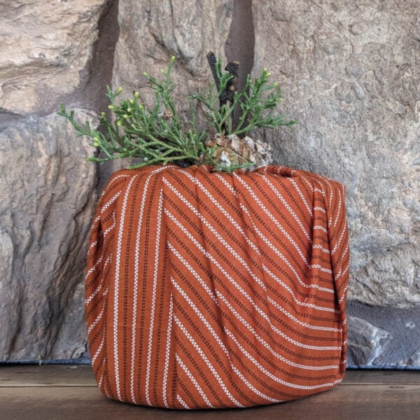 A fabric pumpkin adorned in striped orange textile with greenery at the stem, rests on a wooden surface against a textured stone wall background, perfectly capturing the essence of quilt block artistry.