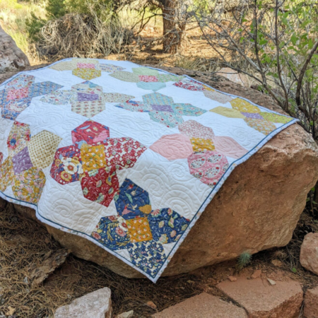 A colorful charm pack quilt with various patterns is draped over a large rock outdoors. Surrounding the rock are smaller rocks and sparse vegetation, creating a natural and rustic setting.