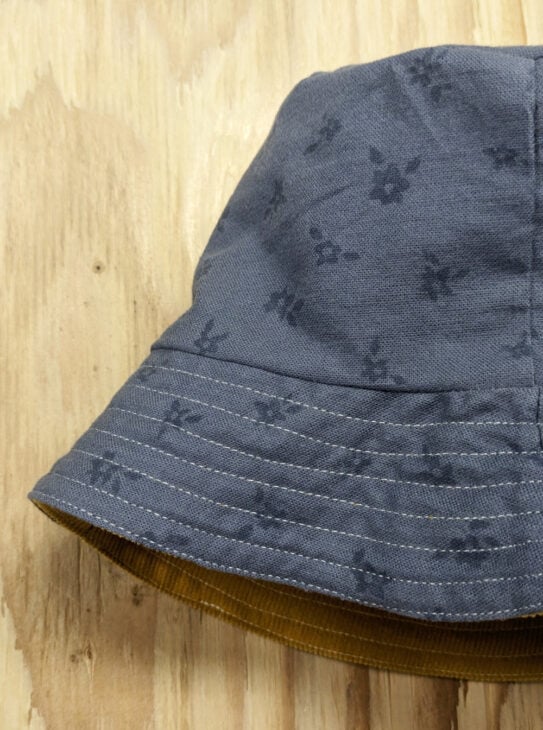 How to Sew a Bucket Hat and it's Reversible! 2 A reversible bucket hat with a subtle floral pattern hangs on a wooden surface. The brim is lined with contrasting yellow fabric inside, adding a pop of color to the thoughtful design.