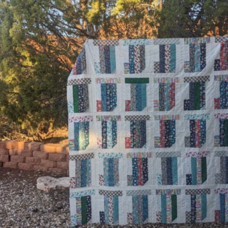 A colorful quilt featuring vertical patterns is displayed outdoors. It hangs in a garden area with a brick wall and green trees in the background, illuminated by warm, golden sunlight, showcasing its Jelly Roll Friendly design.