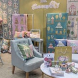 At the Quilt Market 2024, a cozy booth displays colorful quilts and pillows with house and geometric patterns. Soft chairs and tables hold quilting supplies, while decorative wall hangings add charm. A bouquet of flowers sits on a table against a yellow wall backdrop.