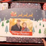Hands hold a festive Christmas Box featuring a card with a couple's photo. Adorned with "Merry Christmas," the scene showcases snowy pines and a sleigh. Filled with decorated gifts, it brings to mind the charm of Jenny Doan's Missouri Star Quilt Company. Scissors are visible nearby, ready for more crafting magic.