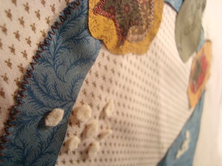 Patchwork Round Robin Border #8 3 Close-up of a quilt showcasing a patterned fabric design. The quilt features blue and yellow floral patches sewn onto a dotted beige background, bordered elegantly. Small white fluffy elements are attached to complete the charming look.