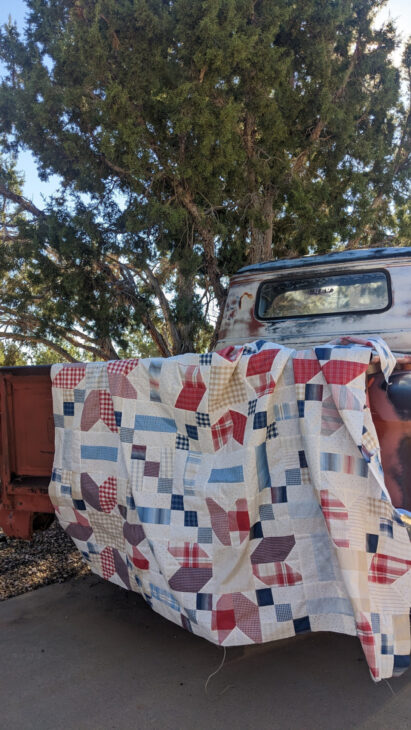 A colorful patchwork quilt with red, blue, and beige patterns is draped over the tailgate of a rusty vintage truck. The truck is parked under a large green tree on a sunny day.