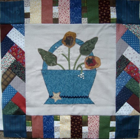 Patchwork Round Robin Border #8 2 A colorful quilt square showcases a central blue basket adorned with flowers and leaves. Encircling it is Border #8—patchwork in various patterns and colors, including red, green, blue, and white—creating a vibrant frame.