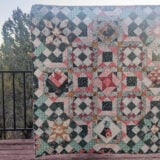 A vibrant sampler quilt, showcasing intricate star patterns and hidden geometric gems, is held up against a wooden deck railing. The backdrop frames a scenic view of trees and a stunning sunset or sunrise sky.