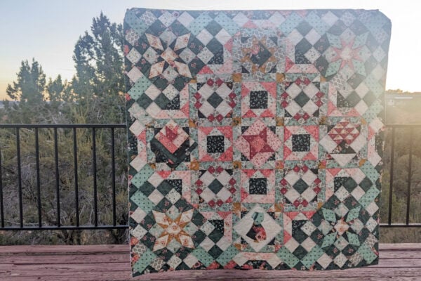 A vibrant sampler quilt, showcasing intricate star patterns and hidden geometric gems, is held up against a wooden deck railing. The backdrop frames a scenic view of trees and a stunning sunset or sunrise sky.