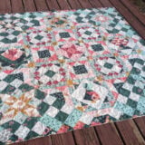 A colorful sampler quilt with intricate geometric and floral patterns in shades of pink, blue, and green is spread out on a wooden deck. This hidden gem features a central star design surrounded by squares and diamonds.
