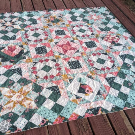 A colorful sampler quilt with intricate geometric and floral patterns in shades of pink, blue, and green is spread out on a wooden deck. This hidden gem features a central star design surrounded by squares and diamonds.