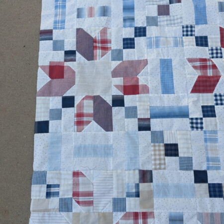 A patchwork quilt featuring geometric patterns in shades of blue, red, and beige, with plaid and striped designs. The quilt is partially spread on a concrete surface.